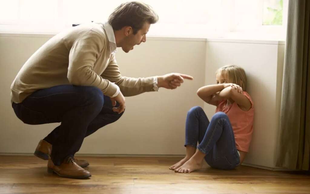 The father is scolding the child, and the child is standing frightened in the corner